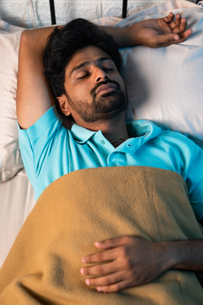 A man with dark hair and a beard is lying in bed on his back, covered with a tan blanket, wearing a teal polo shirt, and resting peacefully with his eyes closed—perhaps dreaming of his new porcelain crowns.