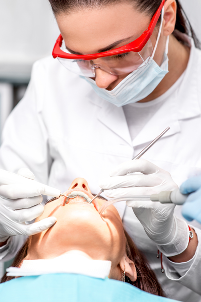 Dentist wearing red safety glasses and a mask uses dental instruments to examine and clean a patient’s teeth, focusing on gum health in a clinical setting.